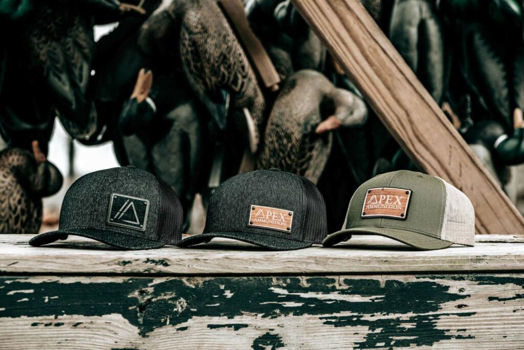 Apex Ammunition branded hunting hats displayed with duck decoys, representing outdoor waterfowl hunting lifestyle gear.